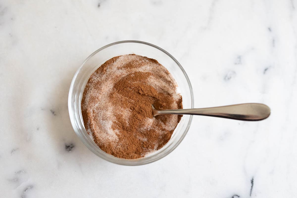 Glass bowl of stirred together cinnamon and sugar with a silver spoon on a white marble worktop.