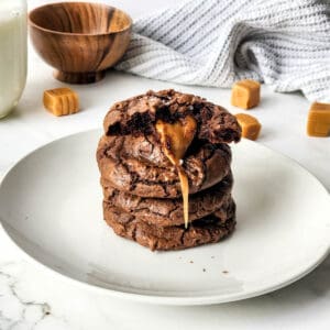 A stack of caramel filled brownie cookies topped with flaky sea salt on a plate with a glass of milk, small bowl, kitchen towel, and caramel candies at the back.