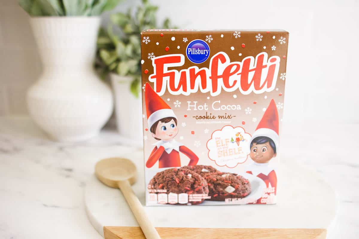 Text on cookie mix box: Funfetti Hot Cocoa Cookie Mix with Elf on the Shelf dolls sitting on the counter next to a wooden spoon.