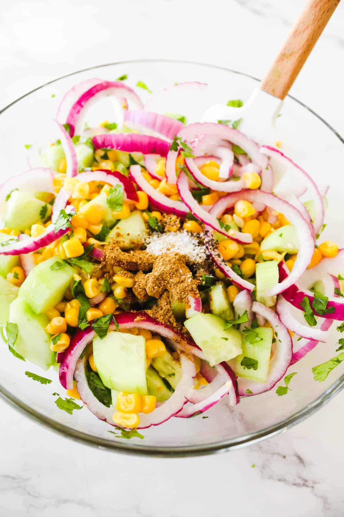 Corn and cucumber salad with red onions, cilantro, cumin, salt, and pepper on a glass bowl with a spatula.