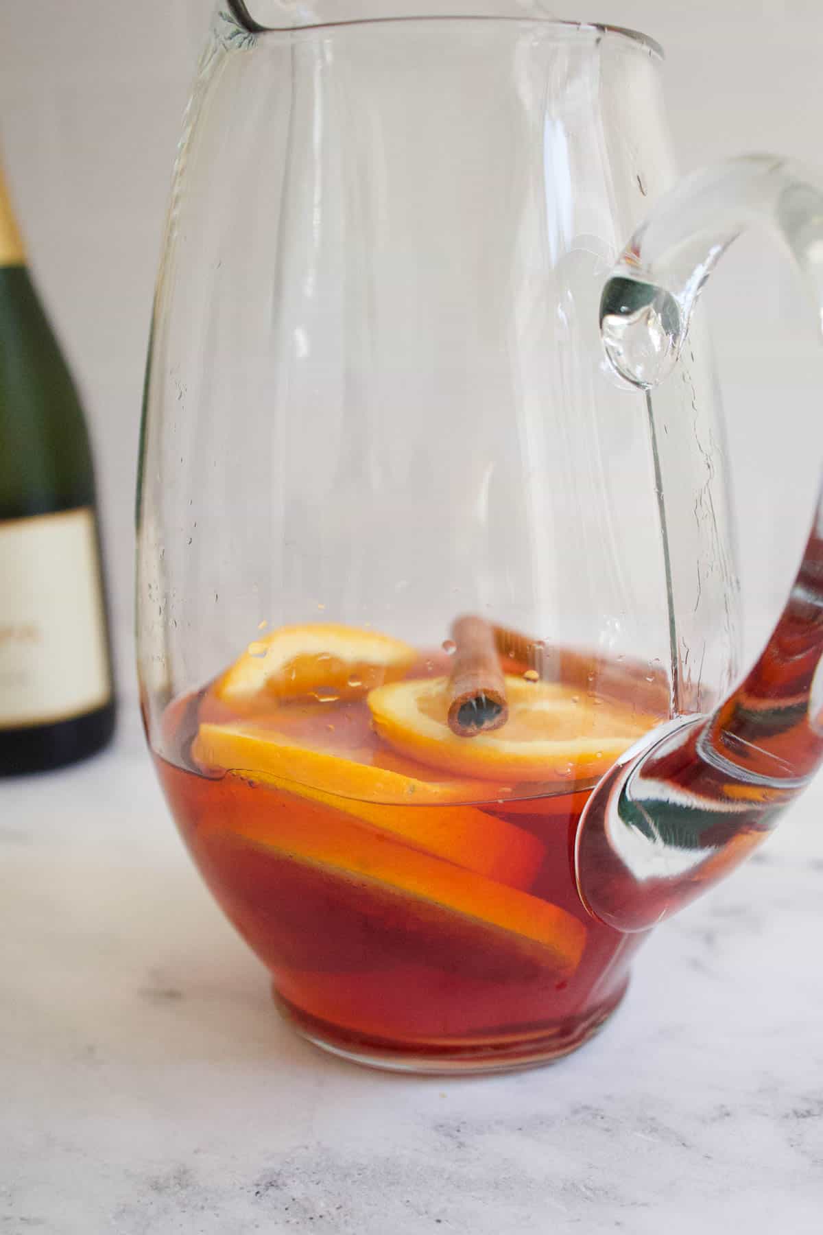 Cranberry juice, sliced oranges and cinnamon sticks in the bottom of a glass pitcher.