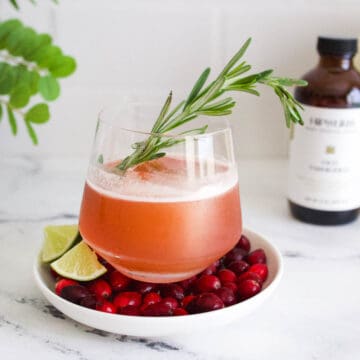 A cranberry old fashioned cocktail garnished with a sprig of rosemary in a glass on top of a plate of fresh cranberries.