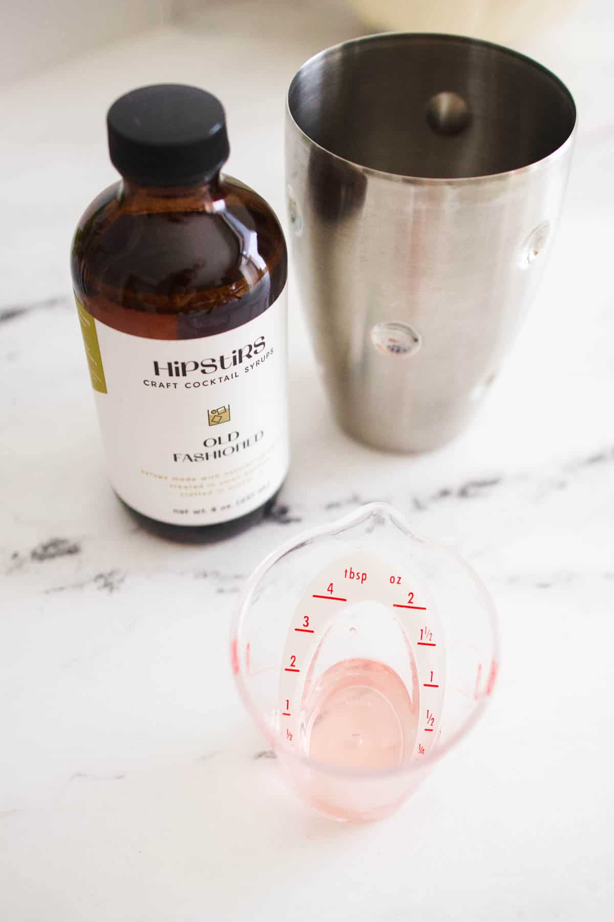 A small measuring cup on a table with cranberry pie simple syrup next to a cocktail shaker.