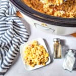 A crockpot on a table with creamy corn inside with a white plate next to it holding a portion of the creamed corn with bacon.