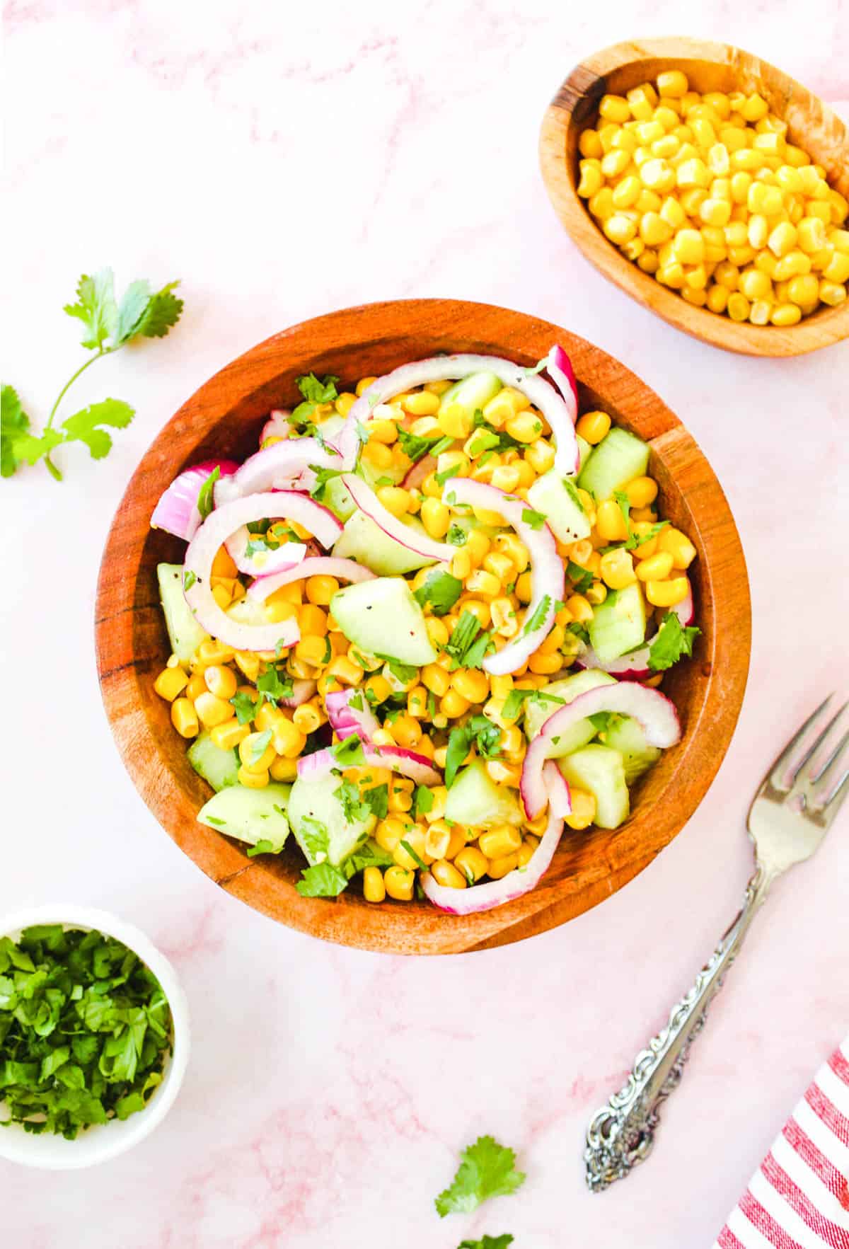 Delicious corn and cucumber salad on a wooden bowl with a fork, cilantro leaves, and a cup of corn kernels on the side.