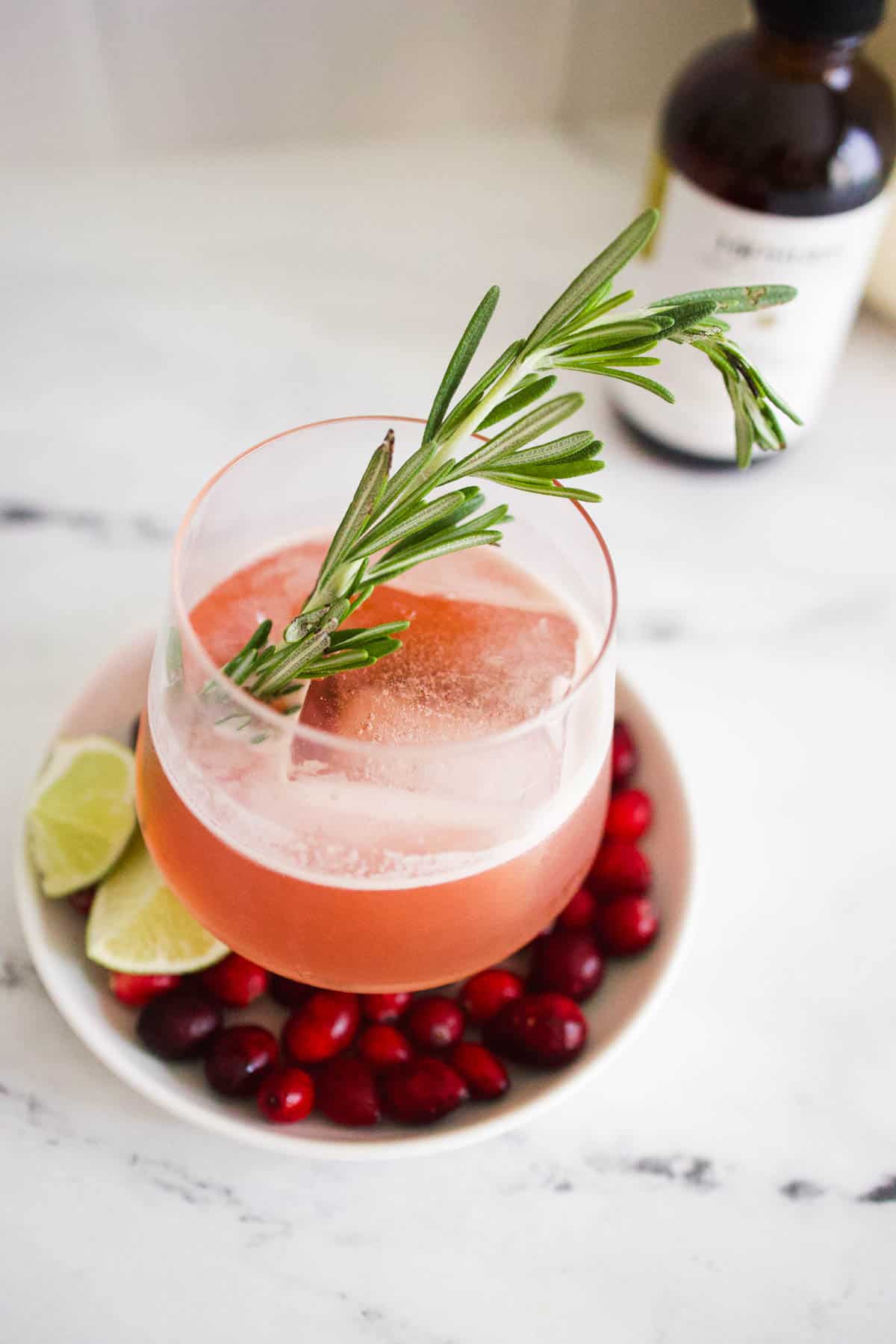 A cranberry old fashioned cocktail sitting on a plate of fresh cranberries with a rosemary garnish.