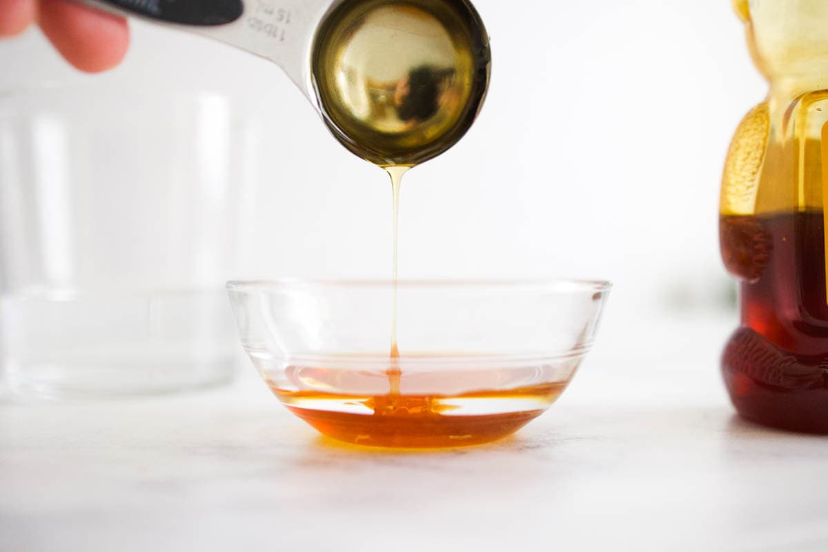 Honey dripping off a measuring spoon into a small glass bowl on a table.