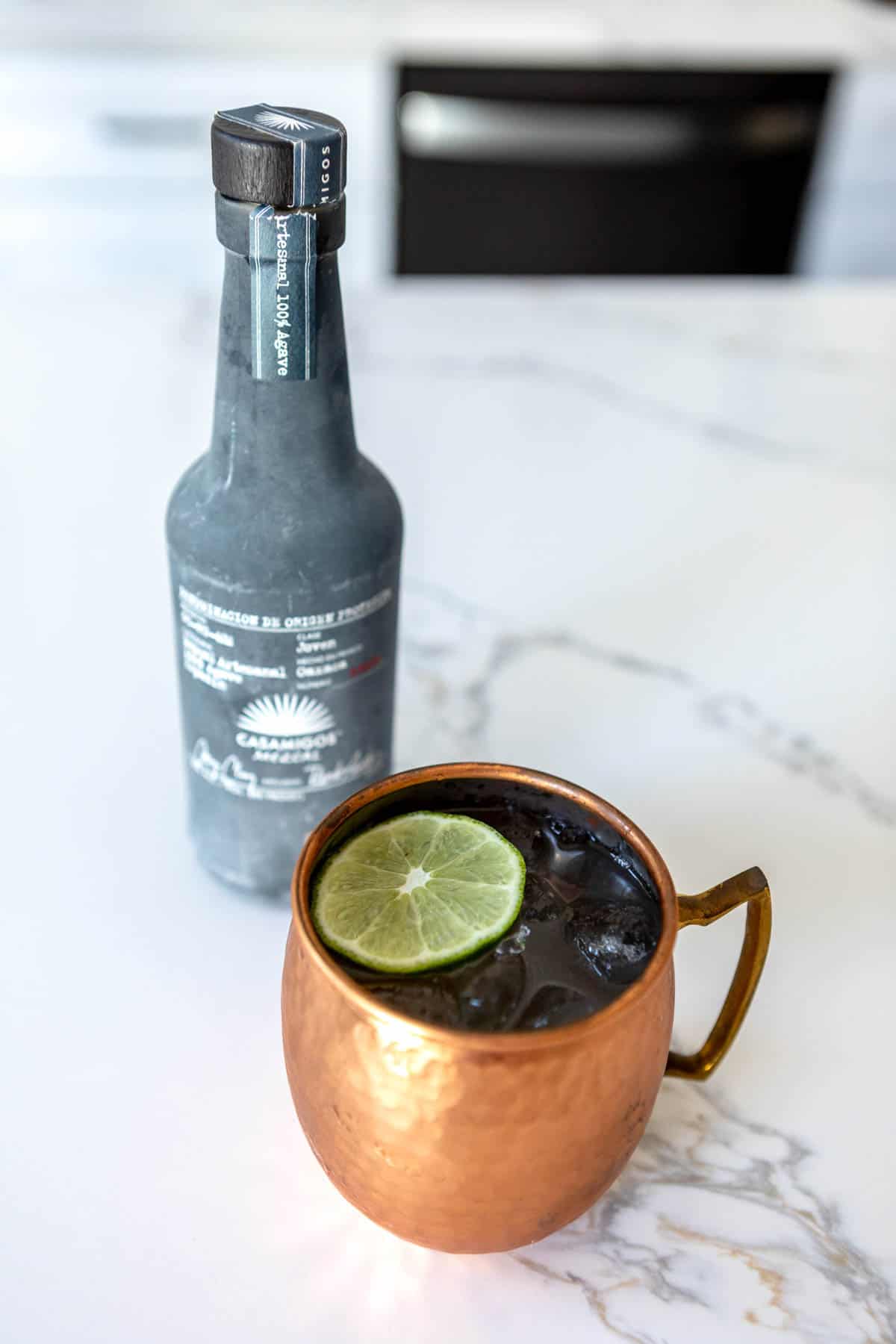 Mule in a copper mug on a white marble countertop next to a bottle of mezcal.