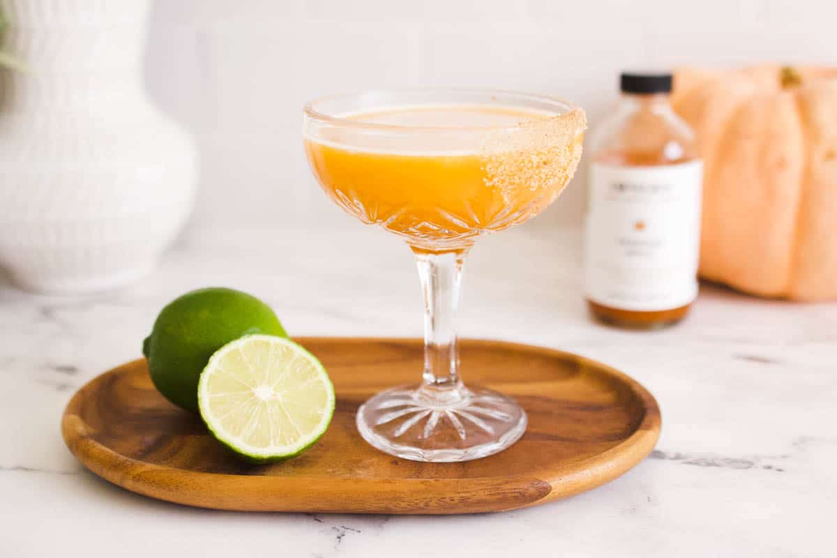 Pumpkin rum cocktail in a coupe glass on a wooden tray with a whole lime and a half a lime.