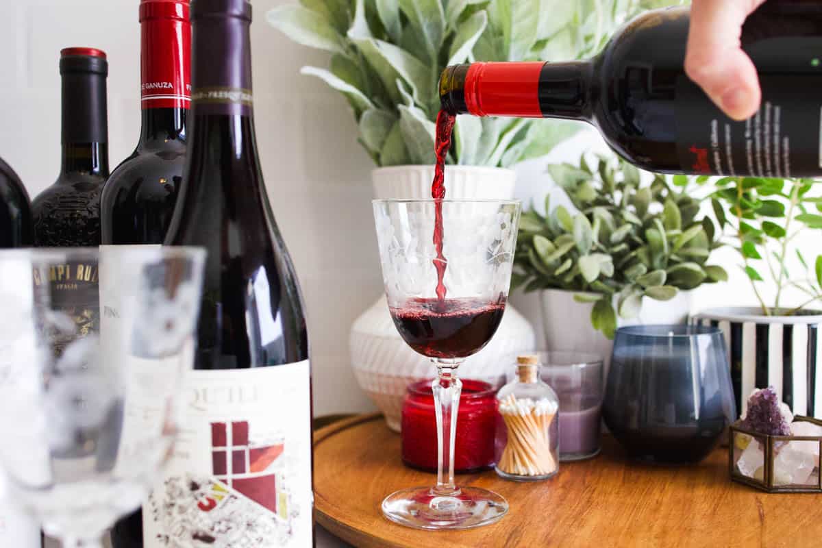 Red wine being poured into a wine glass on a tray next to some bottles of red wine.