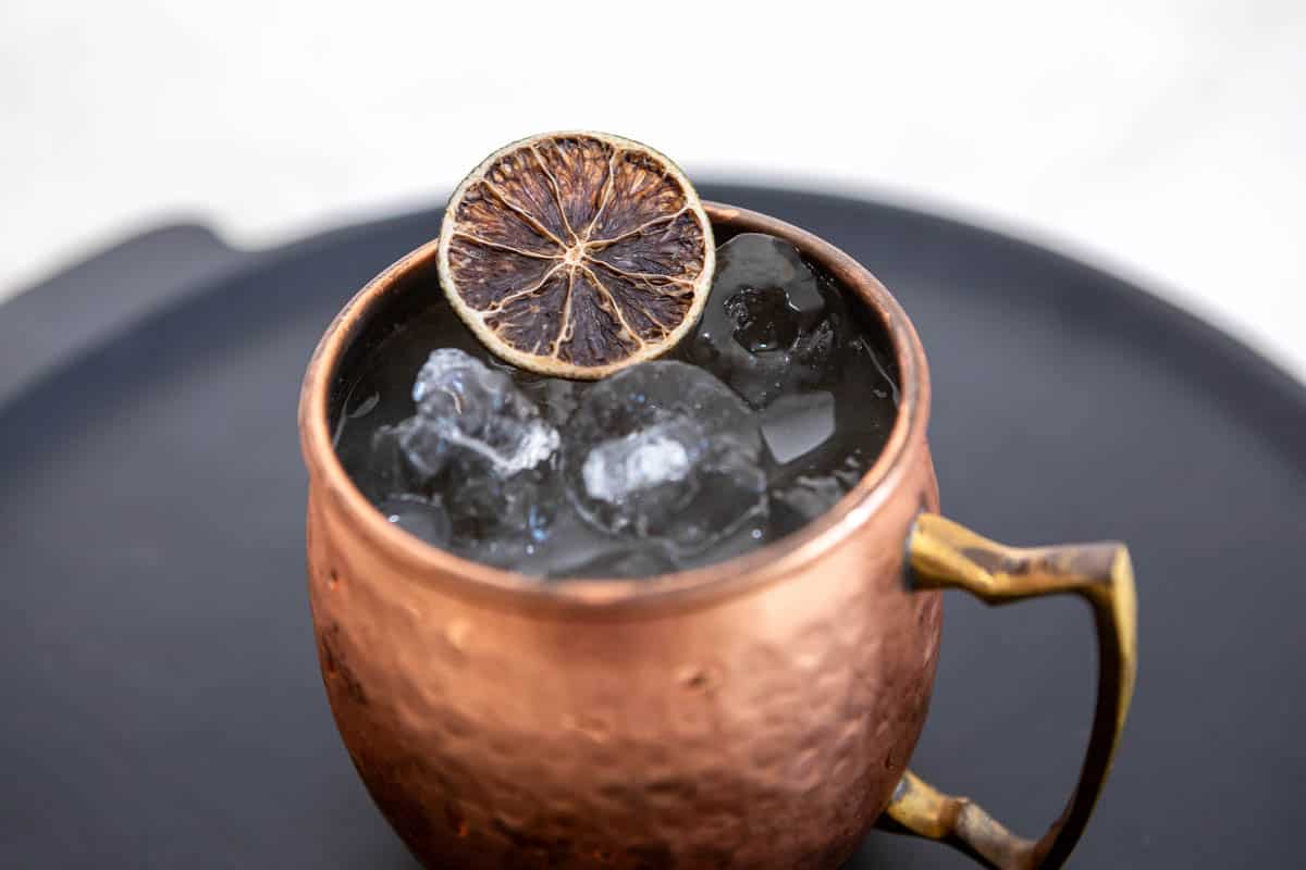 Close up of a copper mule mug with ice and a dried lime wheel garnish on a black background.