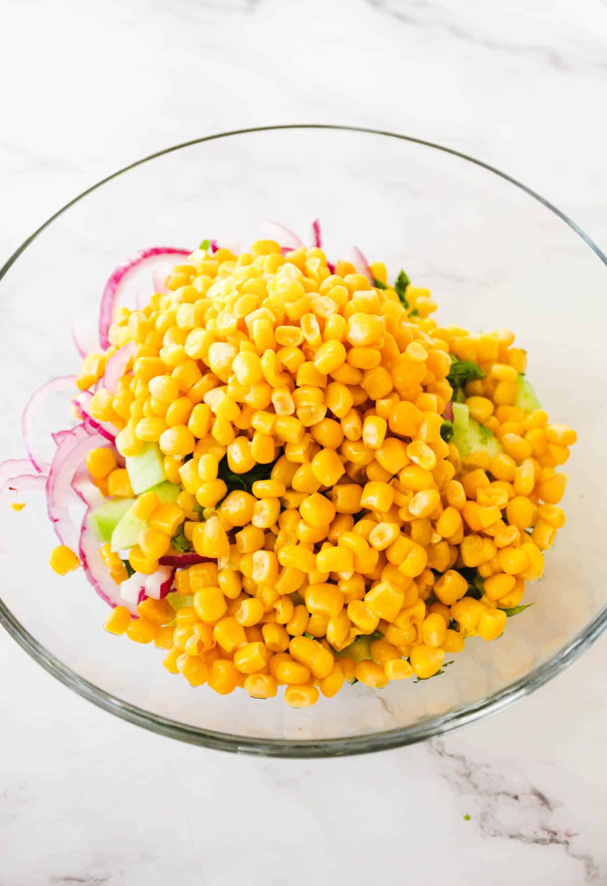 Ingredients needed to make a corn and cucumber salad combined in a glass bowl.