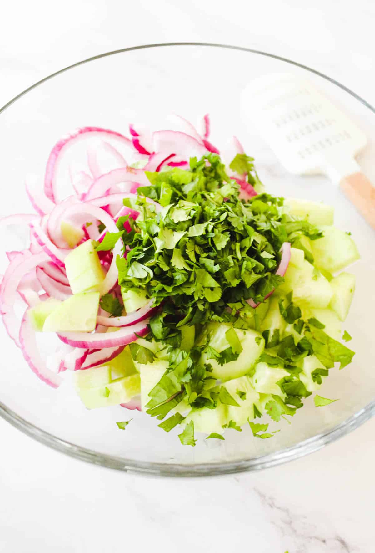 Chopped cucumbers, cilantro, and red onion on a glass bowl.