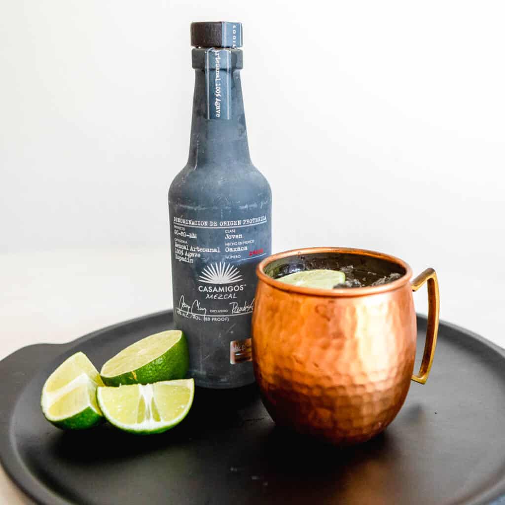 Mule in a copper mug standing next to a bottle of Mezcal and fresh lime wedges.