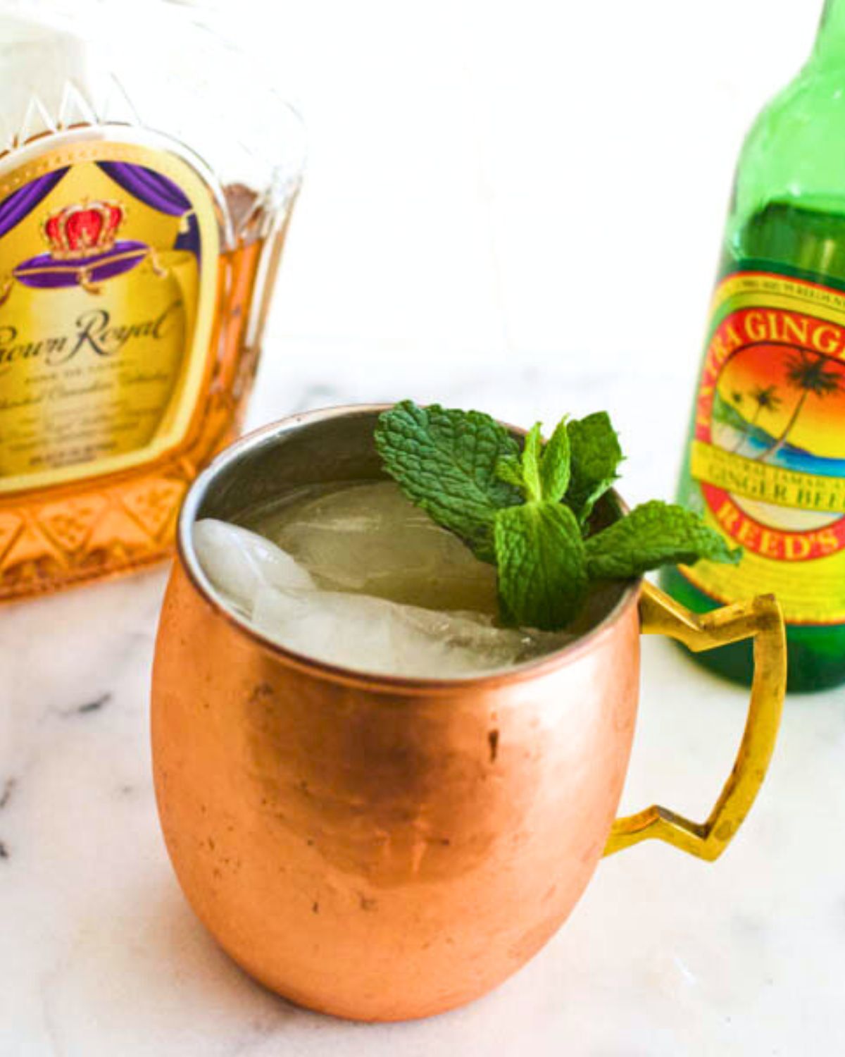 Copper mug holding a whiskey mule topped with mint on a table next to whiskey and ginger beer.
