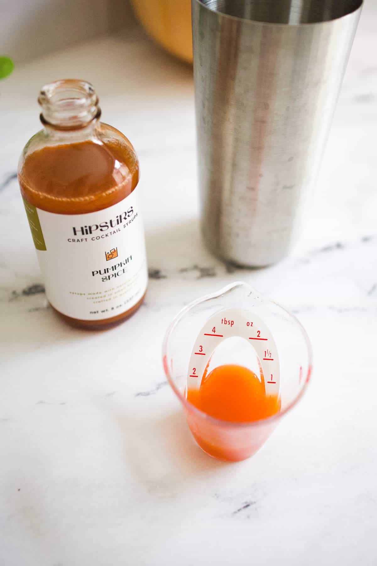 A small measuring cup with pumpkin spice syrup on a table next to a bottle of Hipstirs pumpkin spice syrup and cocktail shaker.