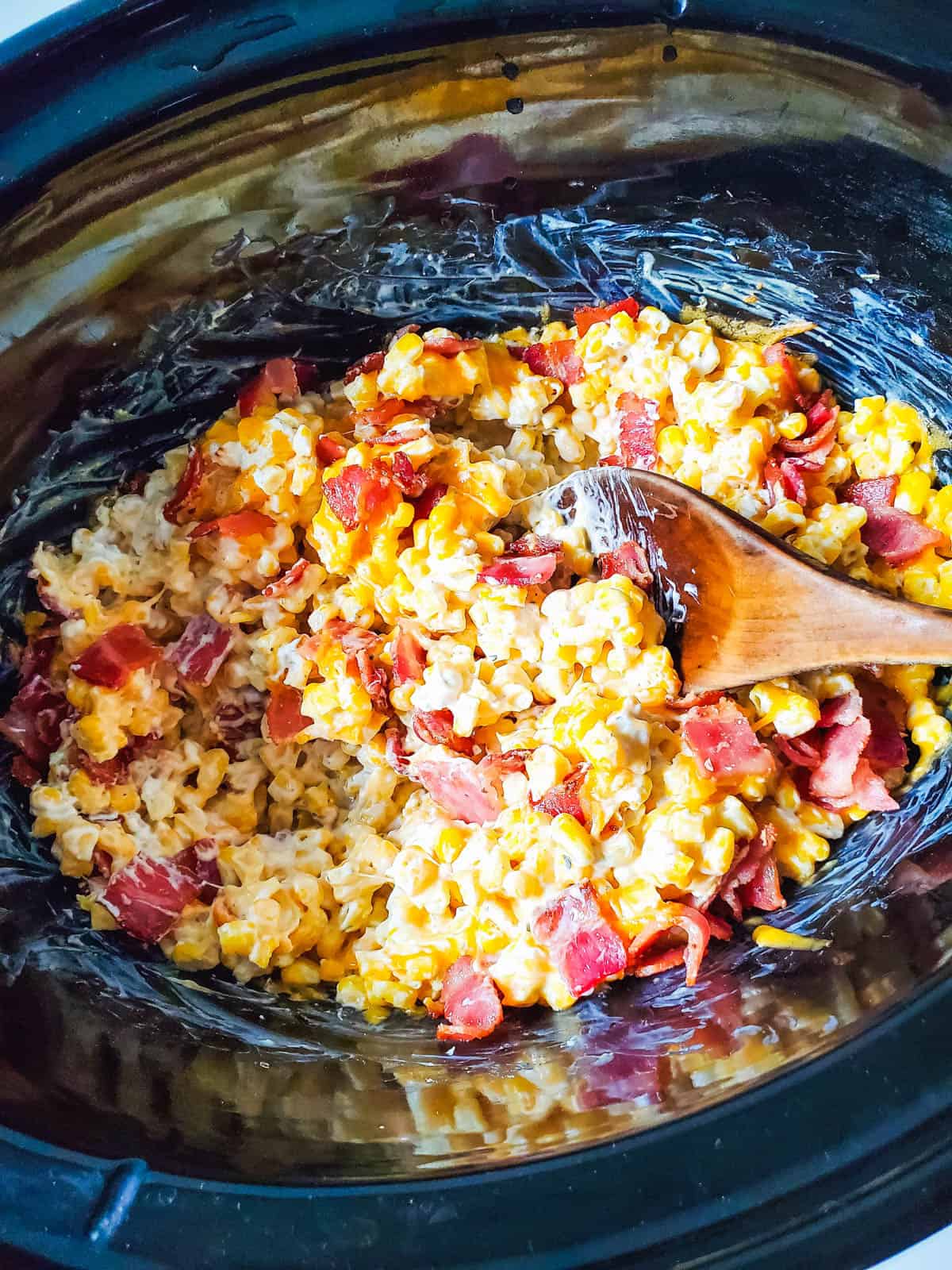 A slow cooker with creamy corn topped with bacon and a spoon stuck in.