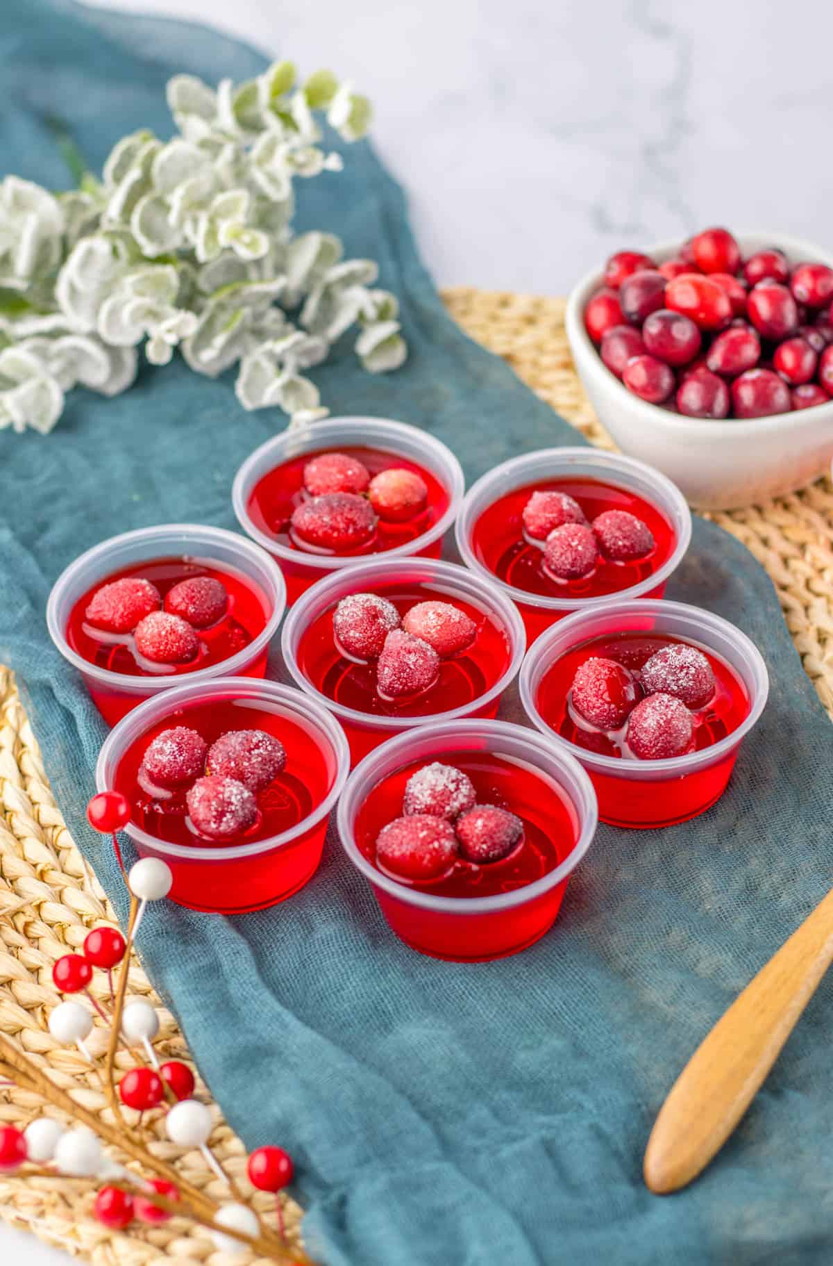 Cranberry Jello shots topped with sugared cranberries on a blue towel next to a bowl of cranberries.