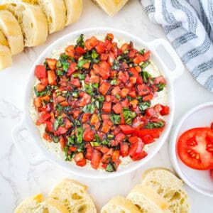 Bruschetta dip in a bowl with a plate of sliced tomatoes and sliced baguette around it.