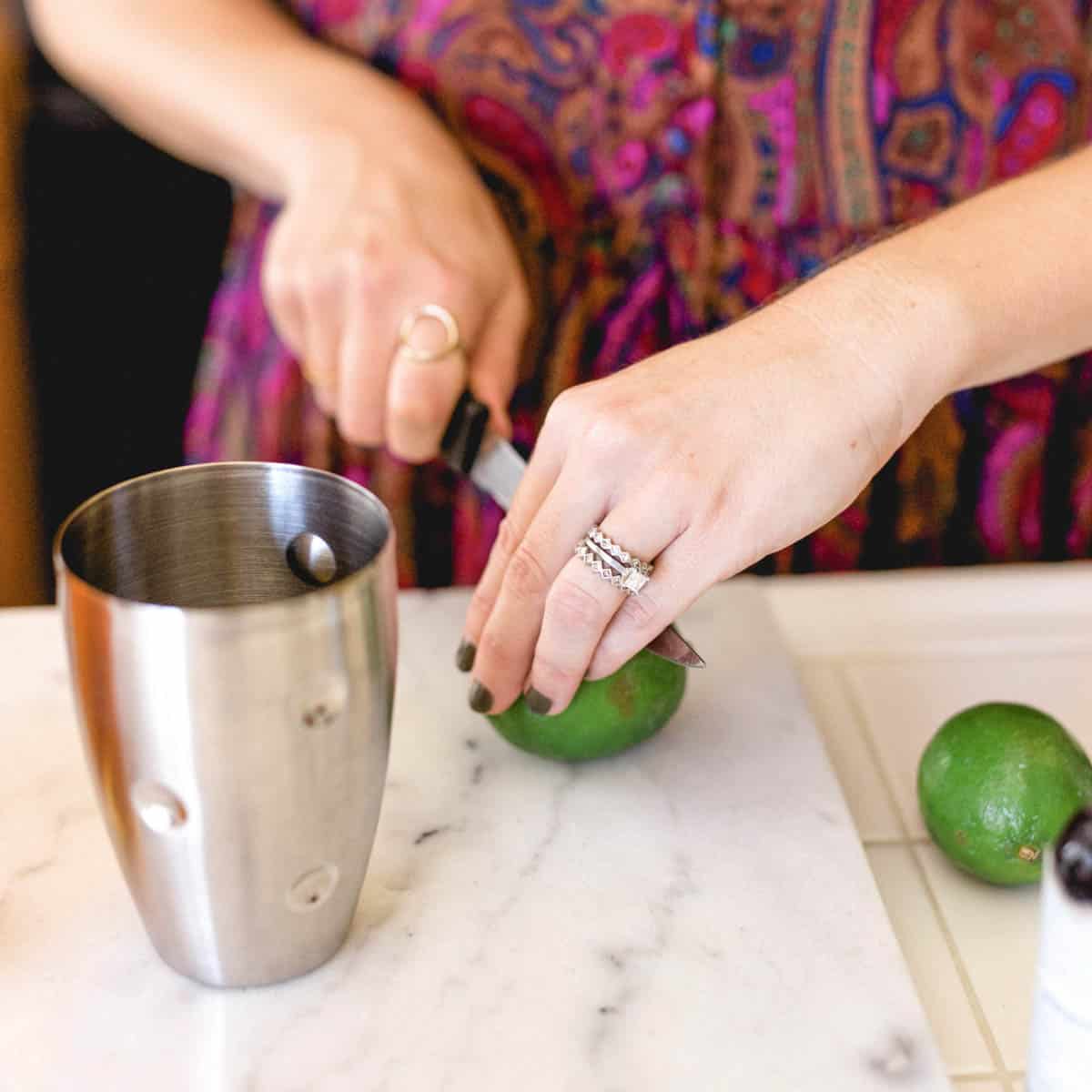Woman cutting a lime on a counter next to a cocktail shaker.