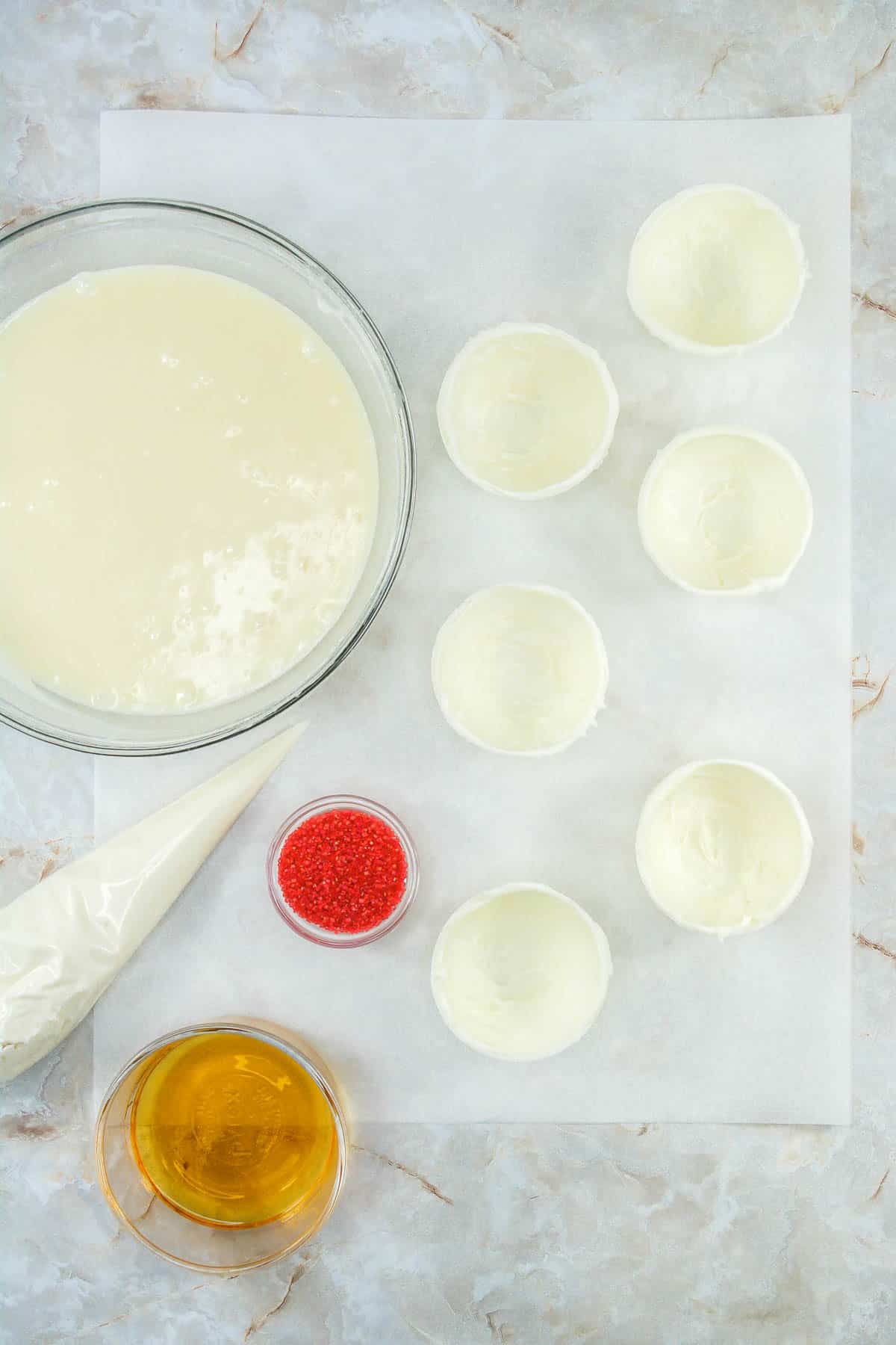 Half spheres of white chocolate next to a bowl of ganache, a piping bag, bowl of whiskey and a small bowl of red sanding sugar.