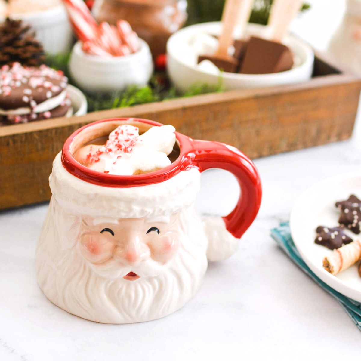 Santa face mug with hot chocolate on a table sitting in front of a tray with toppings to serve on top.