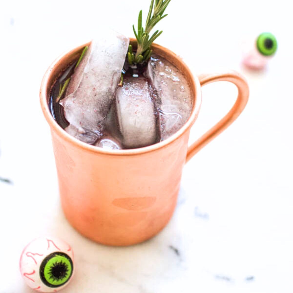 Copper mule mug with ice and a rosemary sprig in a dark colored cocktail with fake eyeballs on the table next to it.