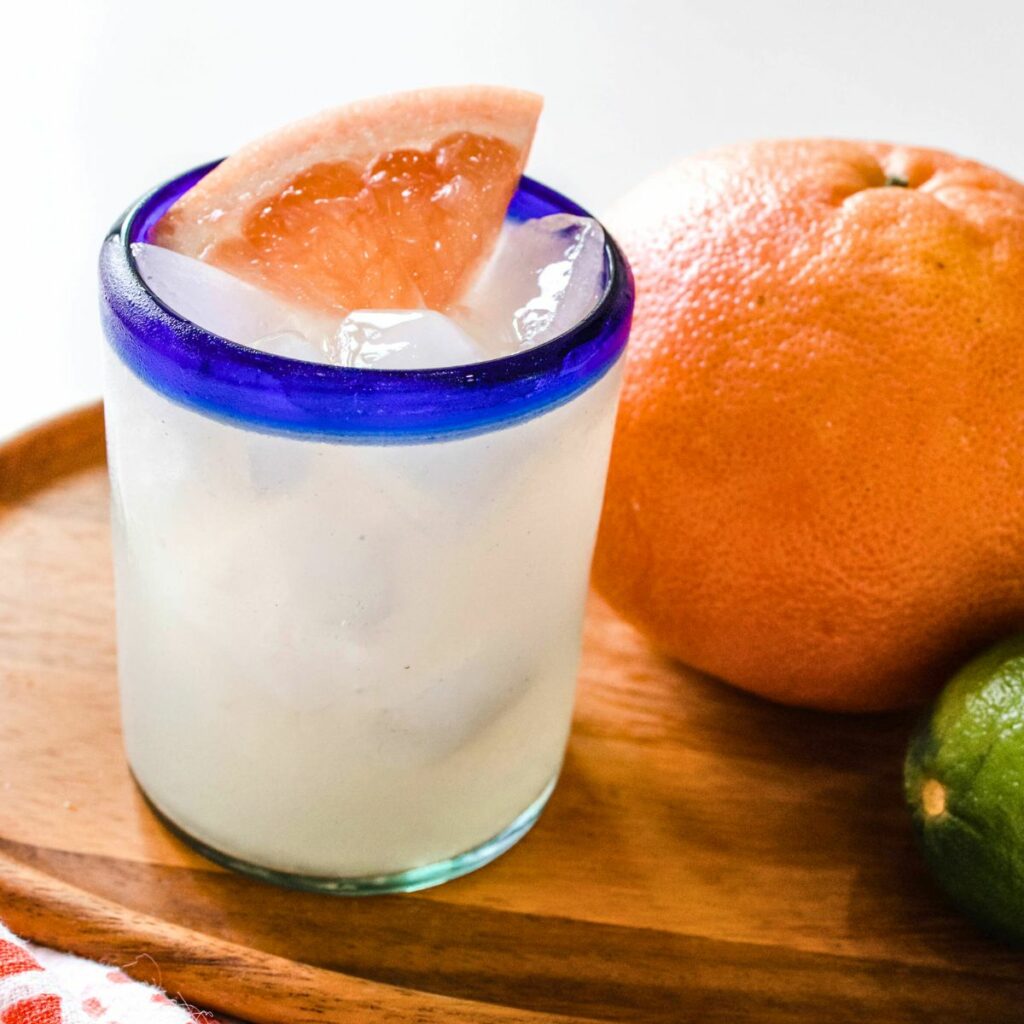 A paloma made with grapefruit juice in a blue rimmed glass topped with pieces of grapefruit.
